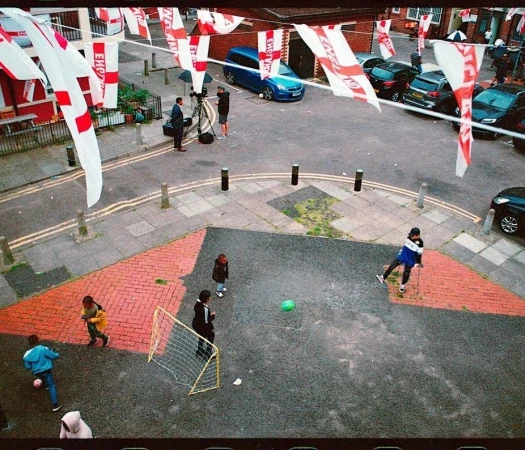 Children play soccer on a street decorated with flags. A news crew films them, with parked cars and buildings in the background.