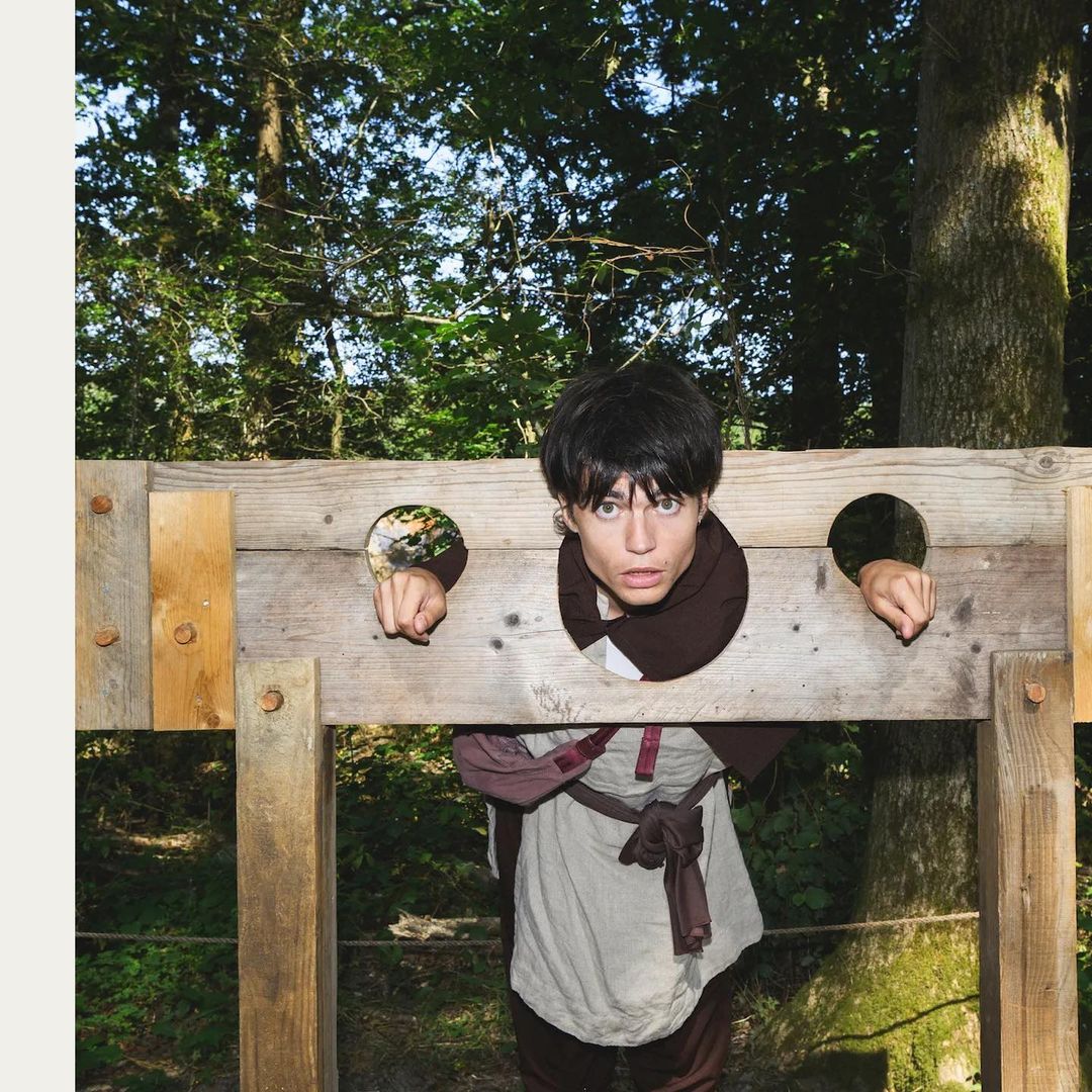 Young man with dark hair in a medieval costume smiles while standing in wooden stocks in a forest with light filtering through the trees.