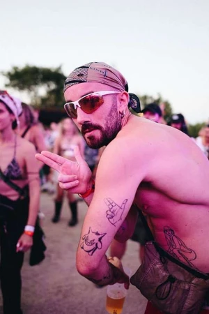Shirtless man wearing sunglasses and a bandana making a finger gun gesture outdoors