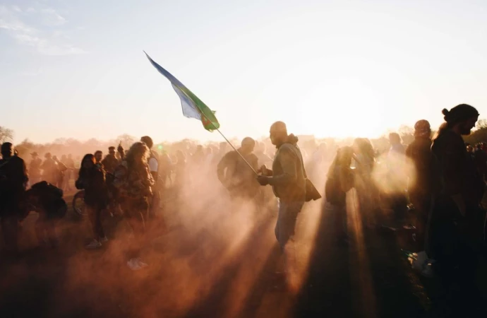 Large crowd of people gathered outdoors, some holding a flag, backlit by sunlight in a wide shot with dust or smoke in the air.