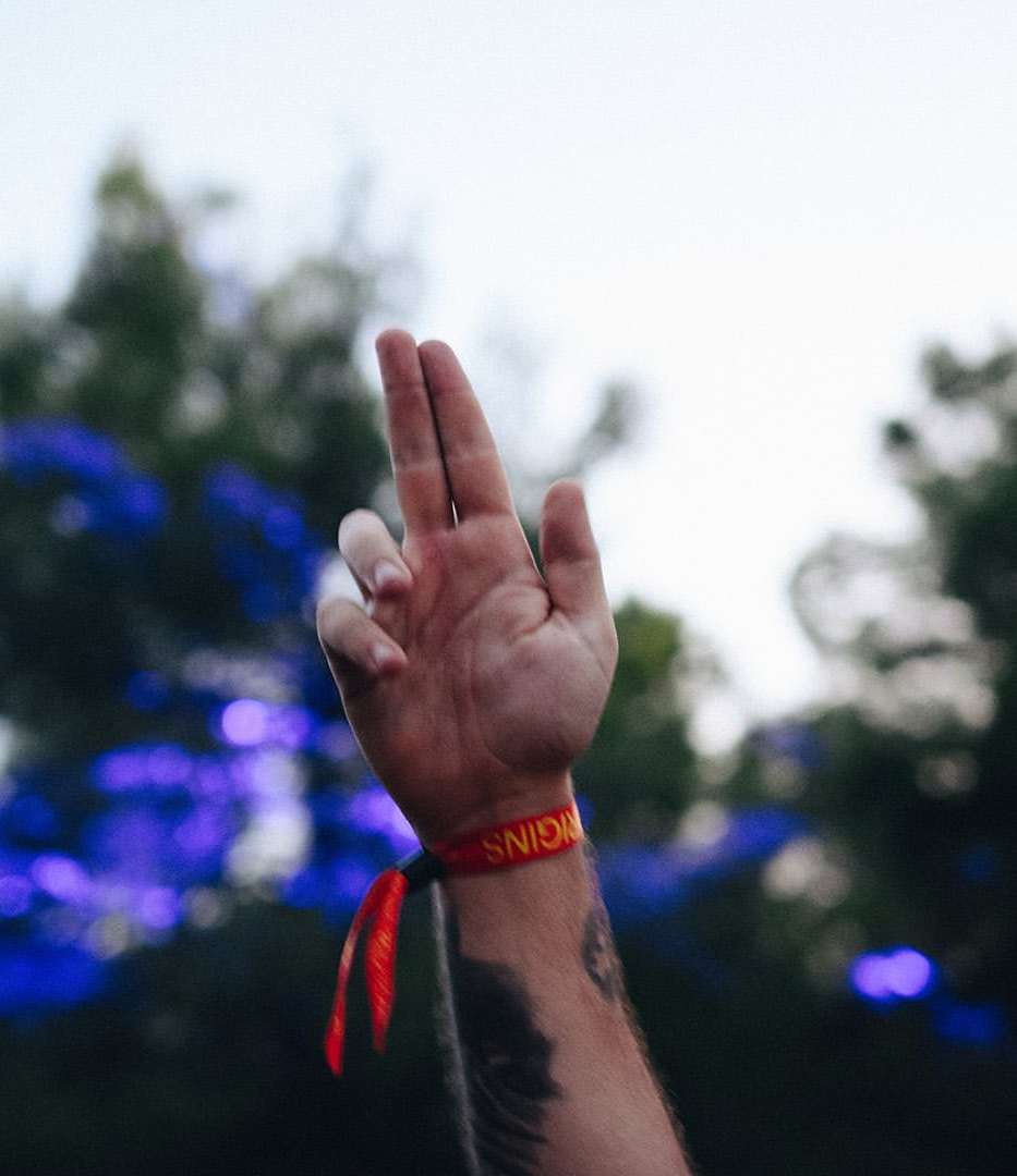 Hand is raised with fingers extended, wearing a red wristband, against a blurry background of blue and green foliage and bright sky.