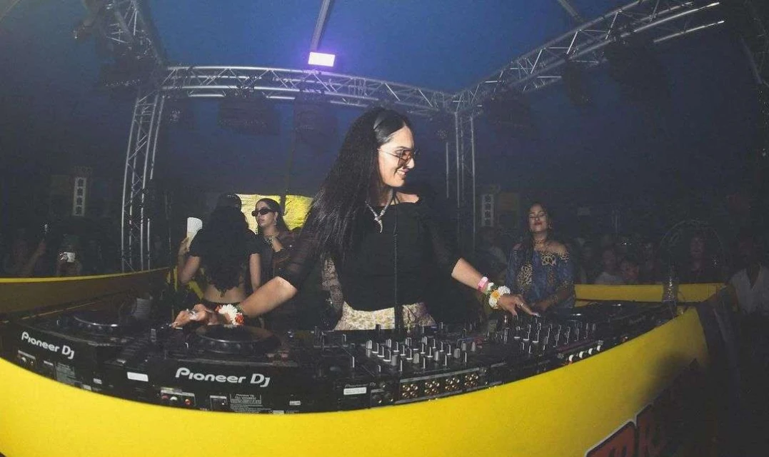 Female DJ performing at a party, wearing sunglasses, black top and gold skirt, with Pioneer DJ equipment and stage lighting.