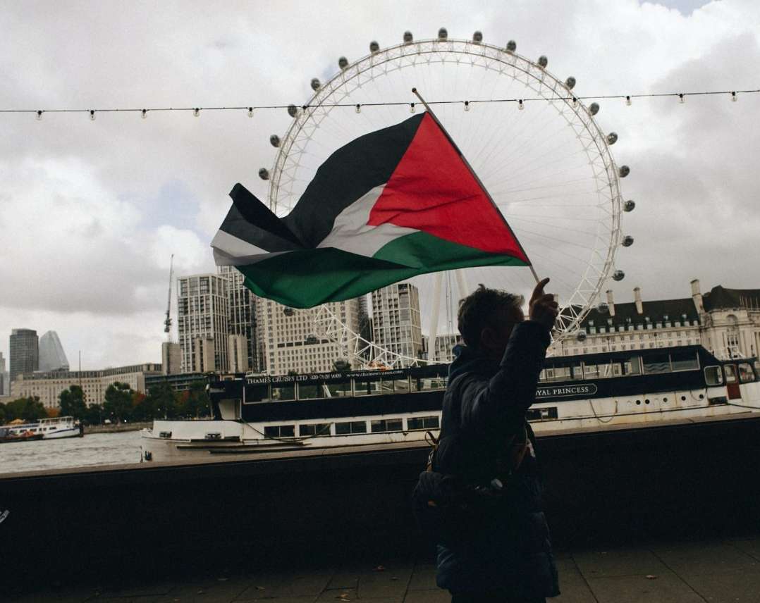 Person holding a Palestinian flag in London with the London Eye in the background on an overcast day in an urban setting.