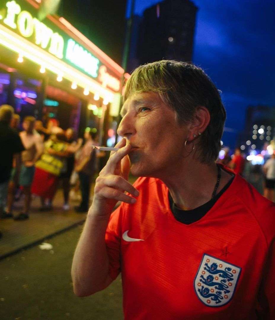 A person in a red jersey smoking outdoors at night, illuminated by building lights with a crowd of people in the background.