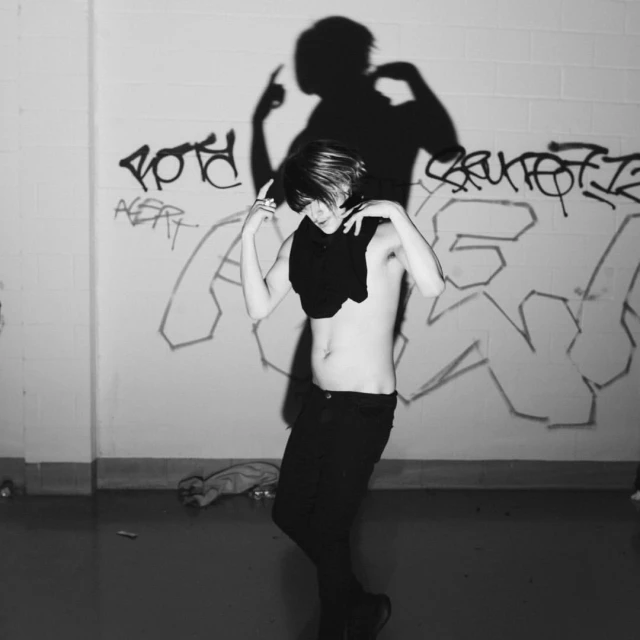 A young adult stands indoors with arms up and hands gesturing near a shadow of a pointing figure against a graffitied wall.