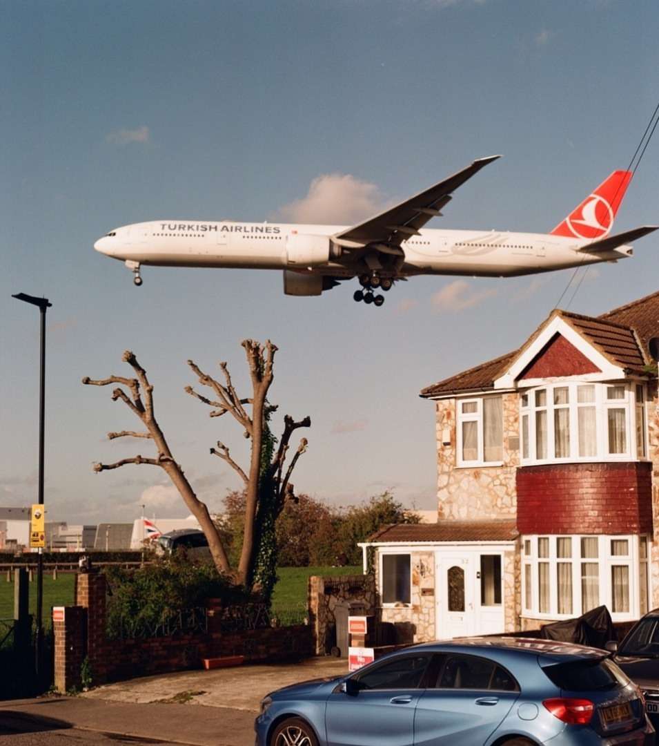 Airplane approaching for landing, flying over a residential area with houses and parked cars under a sunny sky with light clouds.