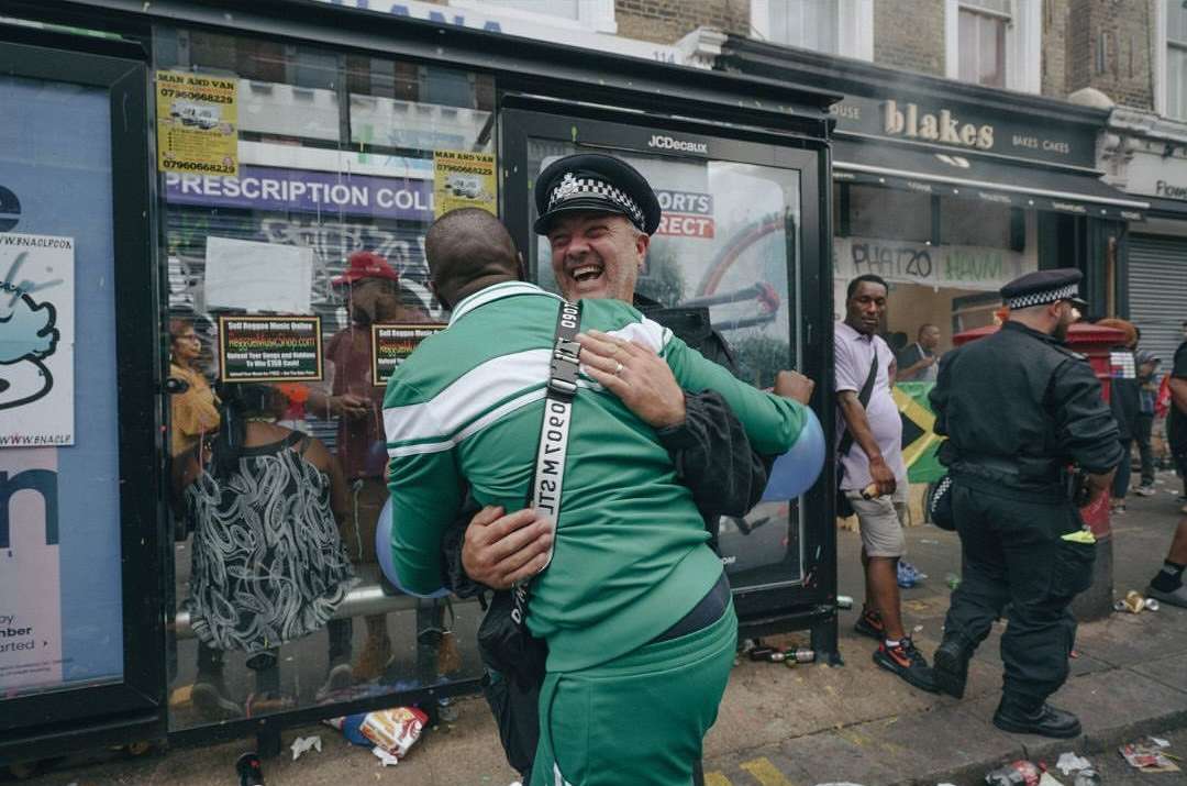 Two men embracing on an urban street with posters in the background; one is a smiling police officer in uniform.