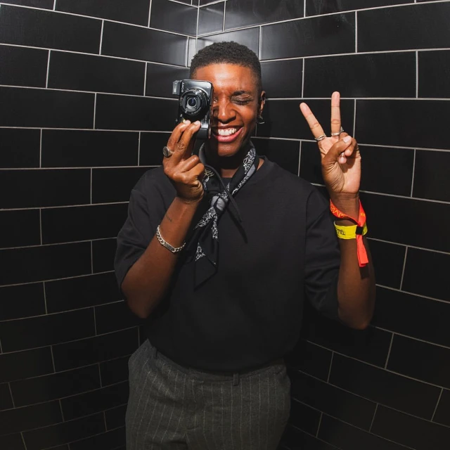 A person holds a camera, smiling and making a peace sign in front of a black tiled wall, creating a candid and engaging portrait.