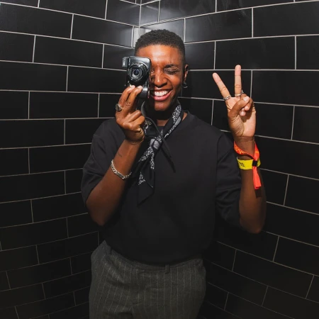 A person holds a camera, smiling and making a peace sign in front of a black tiled wall, creating a candid and engaging portrait.
