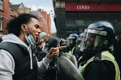 Man pointing at a line of riot police officers wearing helmets and protective gear in a London street by Yushy Pachnanda