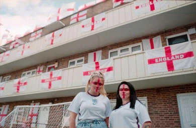 Two women wearing England football shirts and face paint standing in front of a brick apartment building draped with English flags