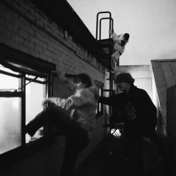 Monochrome image of three young men interacting on a brick building rooftop with an exterior metal staircase