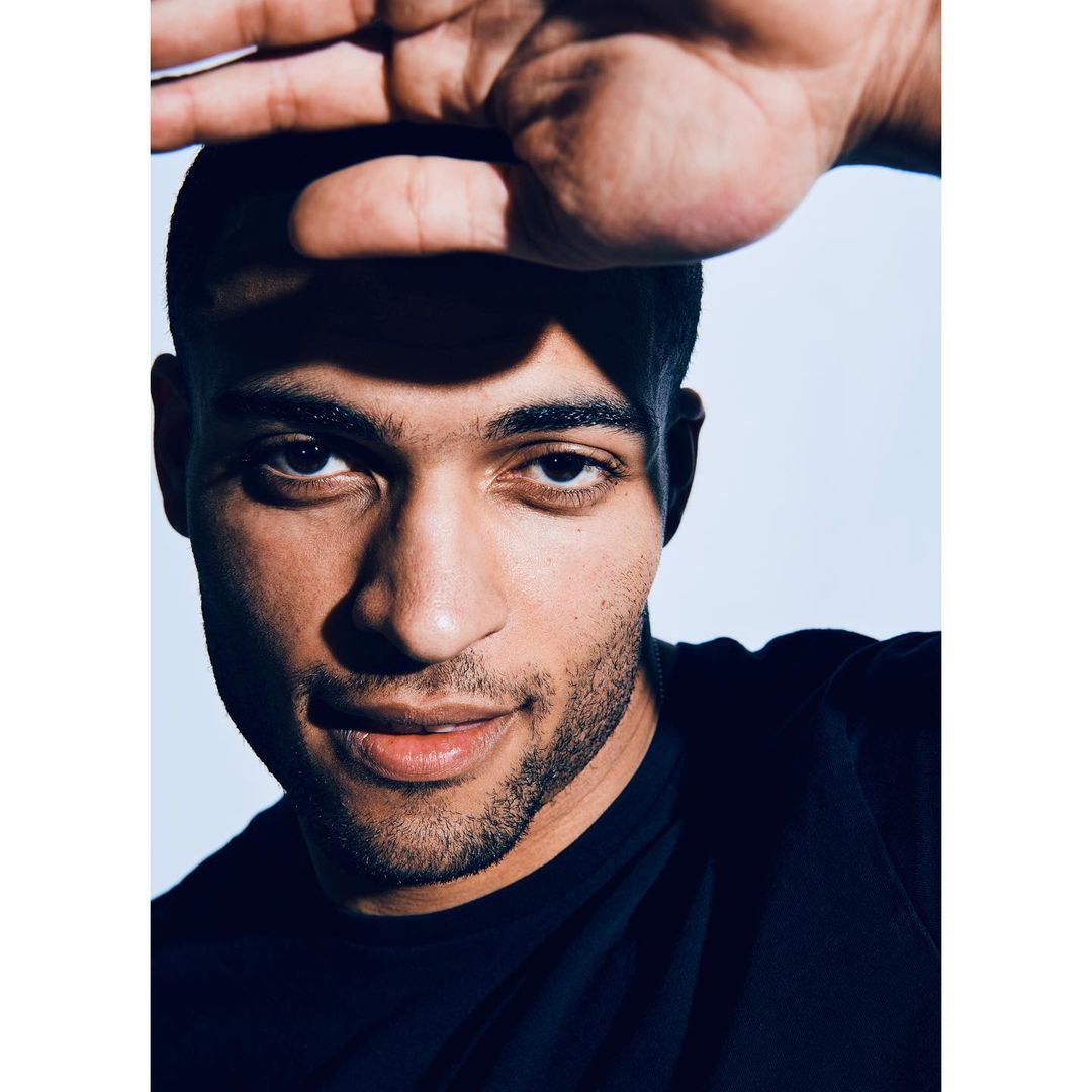 Close-up studio portrait of a young adult male with dark hair, shielding his face from the light with his hand, wearing a black shirt.