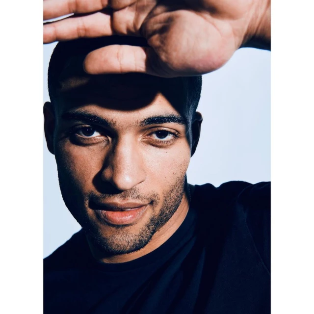Close-up studio portrait of a young adult male with dark hair, shielding his face from the light with his hand, wearing a black shirt.