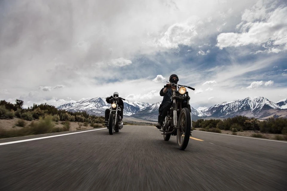 Two motorcyclists ride down a rural highway with snow-capped mountains in the distance under a cloudy sky in a wide, outdoor shot.