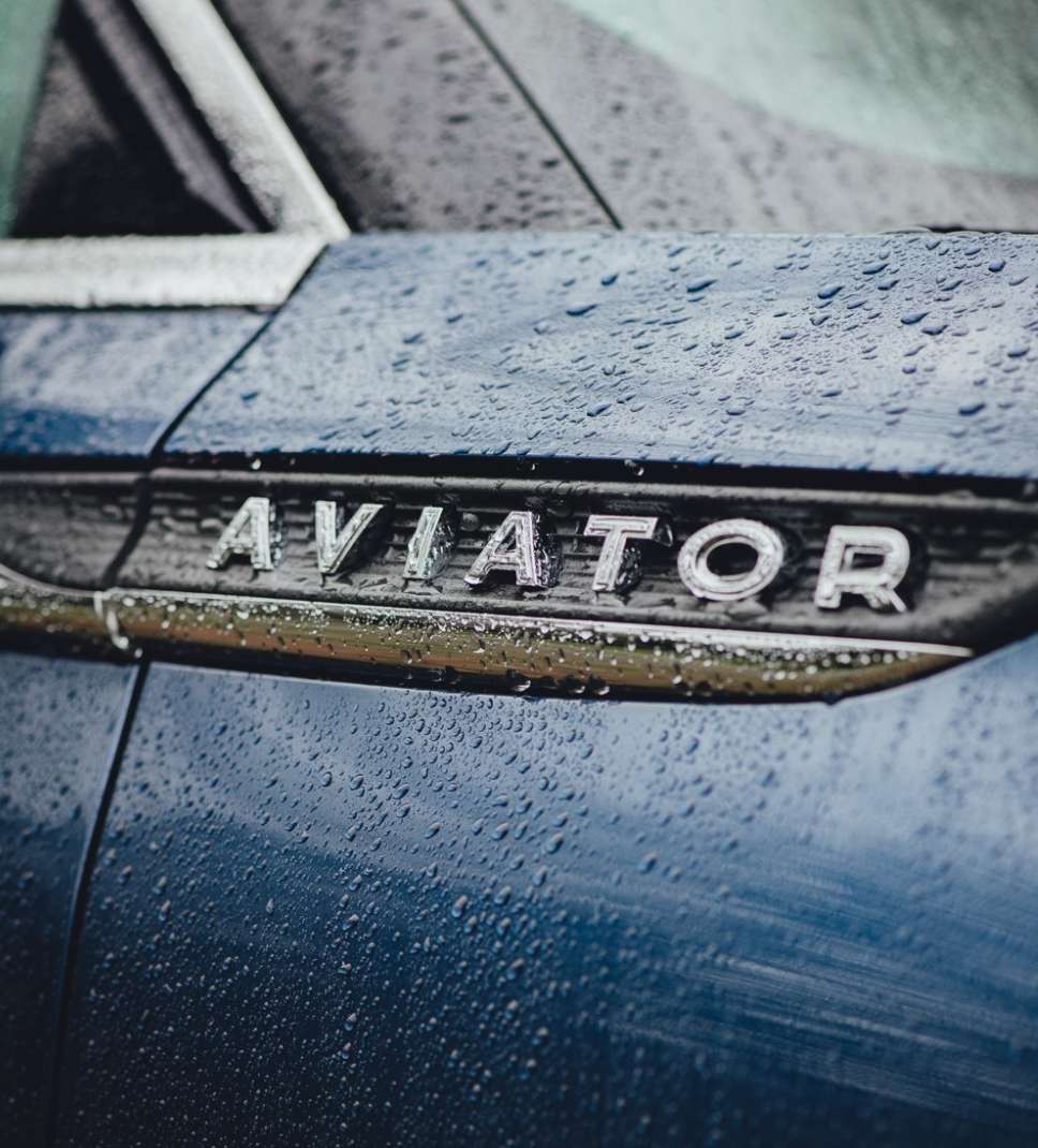 Close-up shot of a blue Lincoln Aviator car with water droplets on the surface, highlighting the chrome 'AVIATOR' emblem.