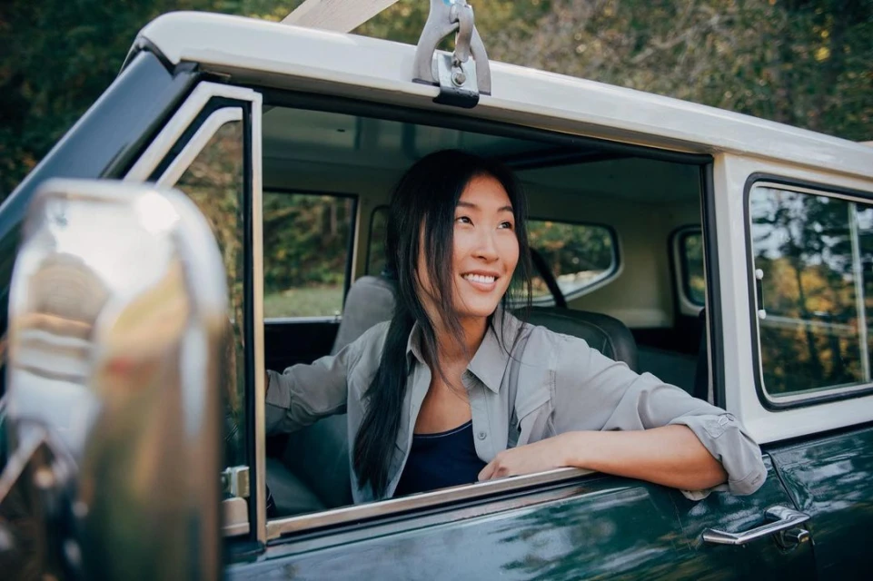 Smiling woman inside a vintage car with a forest backdrop, showcasing automotive design and outdoor lifestyle.