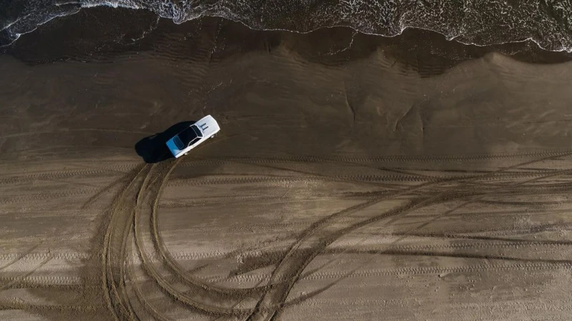 Aerial view of a white car driving on sandy beach leaving tire tracks, with ocean waves in the background, captured in an outdoor setting.