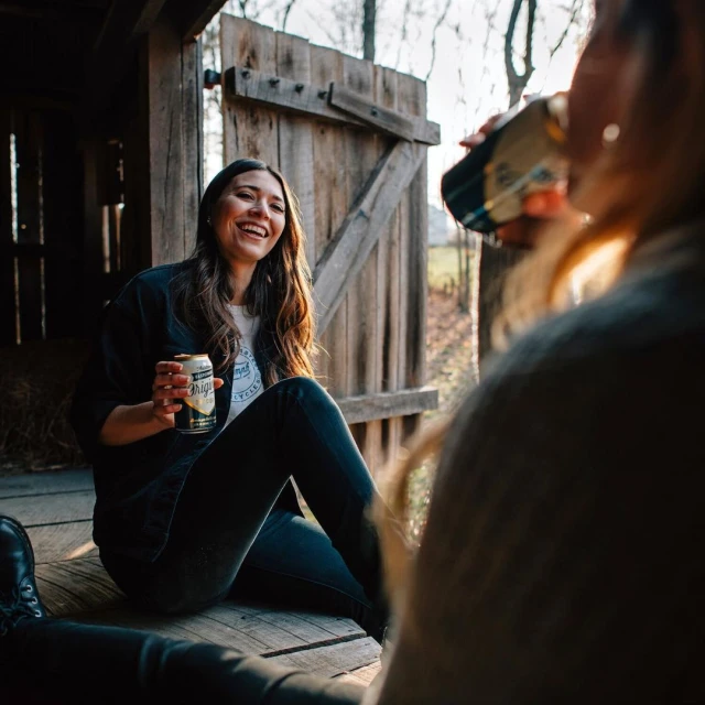 Two women share beers and conversation in a rustic outdoor setting; one woman laughs as she sits on a wooden porch near a shed.