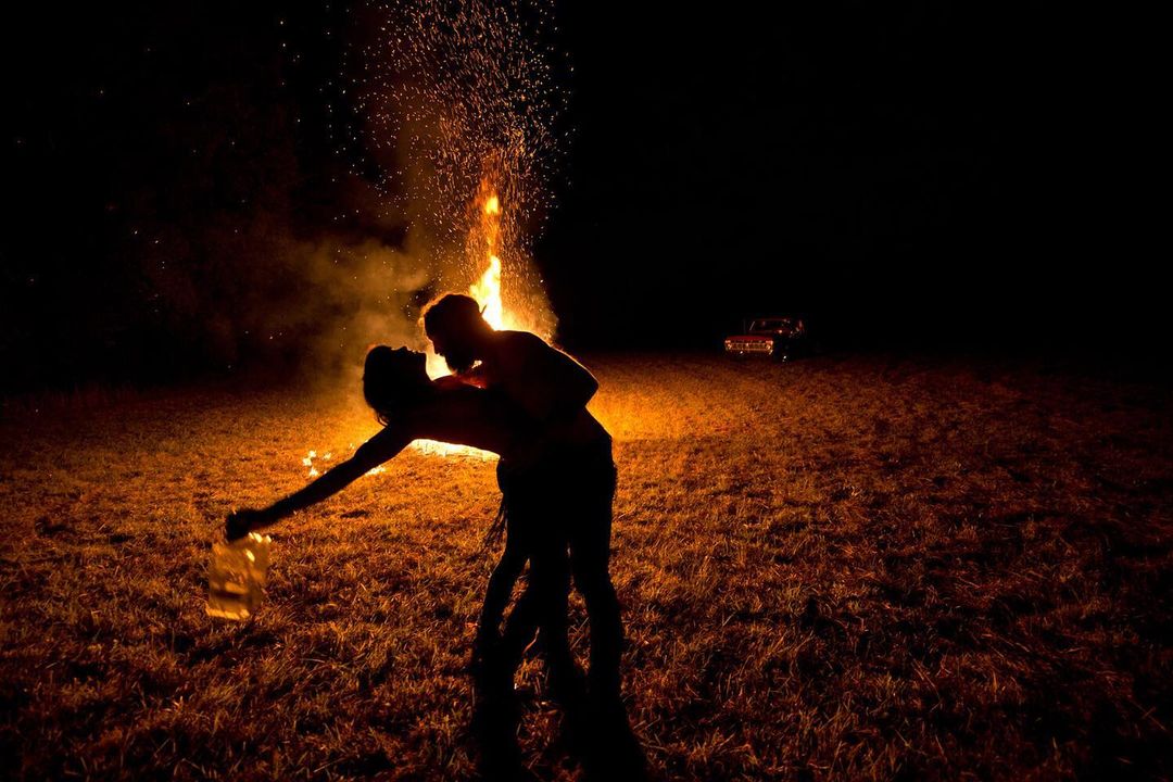 Silhouette of a couple embracing near a bonfire under a night sky, with a classic car parked in the distant background.