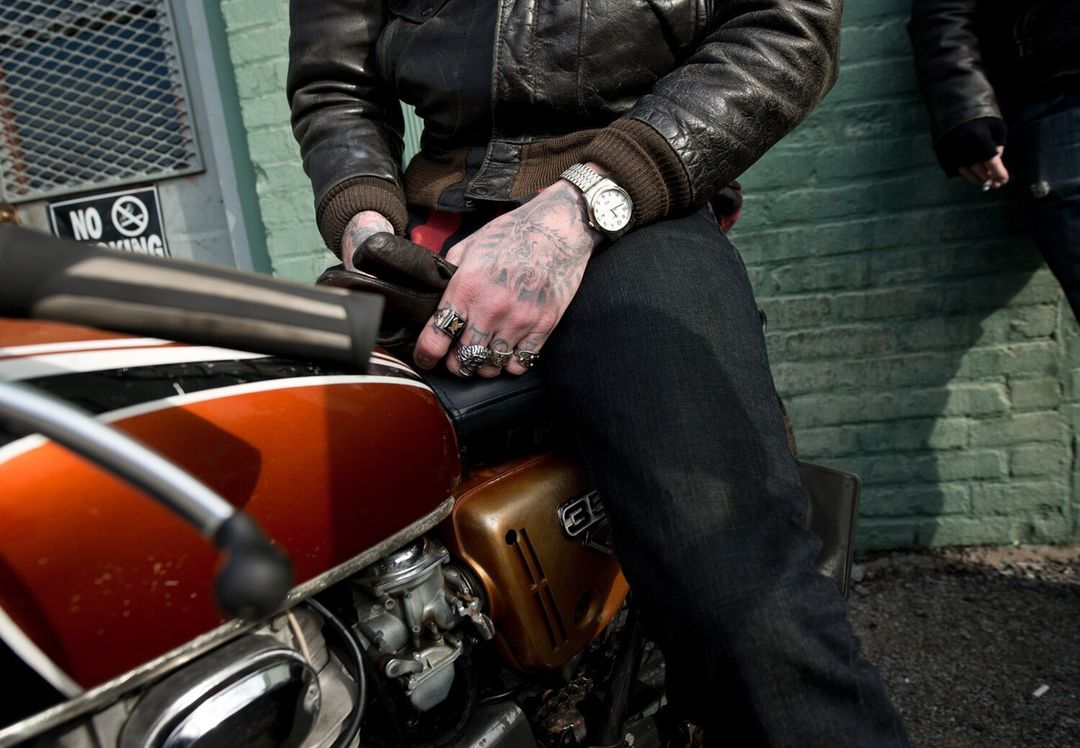 Person sitting on an orange and black motorcycle with tattoos on their hands and wearing a black leather jacket and watch, in an urban setting.