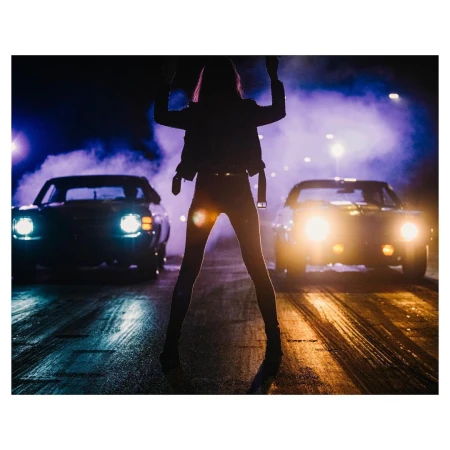 Silhouette of a woman standing on an asphalt surface between two cars with headlights on against a dark, smoky background.
