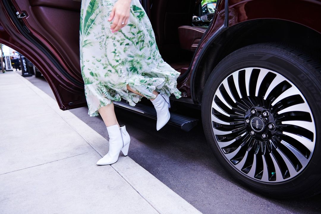 Woman in a floral dress and white boots exiting a luxury car on a city street, captured in bright daylight with shallow depth of field.