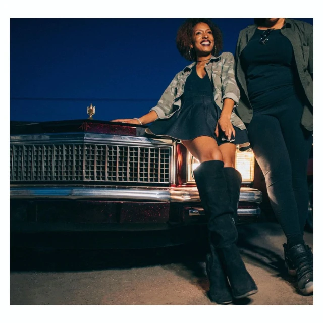Two women pose in front of a classic car at night. One woman leans on the car, wearing boots, a mini skirt, and a camo jacket.