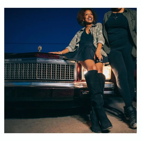 Two women pose in front of a classic car at night. One woman leans on the car, wearing boots, a mini skirt, and a camo jacket.
