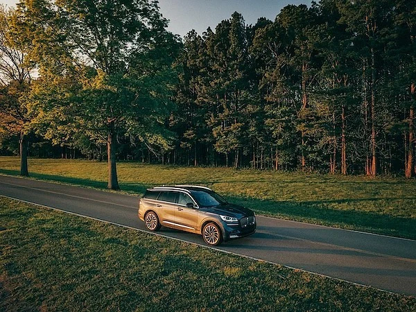 Lincoln Aviator SUV driving on a paved road surrounded by green grass and a lush forest, creating a scenic and natural view.