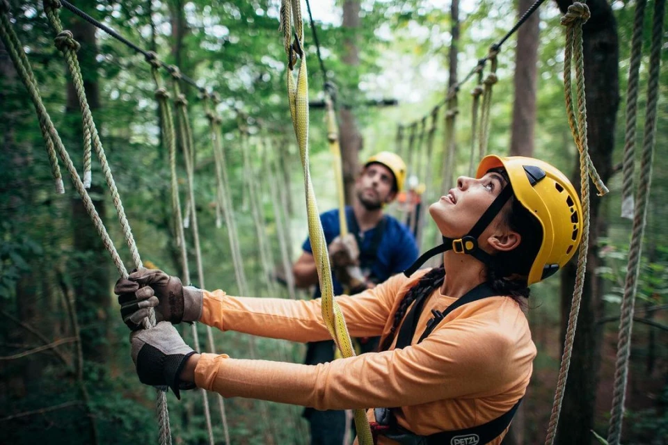 Woman in safety helmet and harness on a ropes course, holding onto ropes while a man looks on in a wooded outdoor environment.