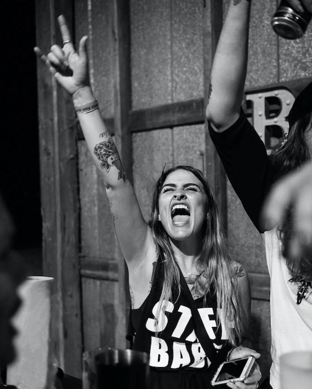 Enthusiastic woman with tattoos cheers with a rock-and-roll hand gesture, holding a smartphone at a concert in a black and white photo.