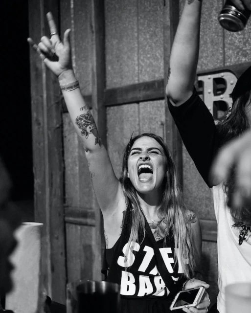 Enthusiastic woman with tattoos cheers with a rock-and-roll hand gesture, holding a smartphone at a concert in a black and white photo.