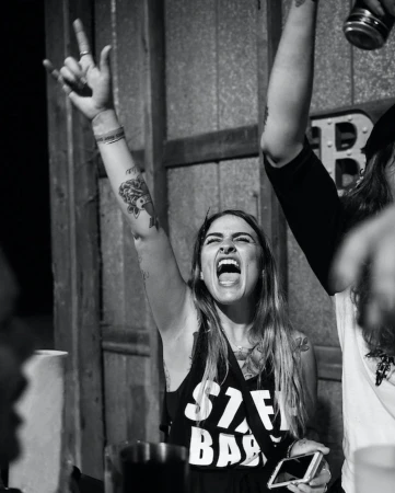 Enthusiastic woman with tattoos cheers with a rock-and-roll hand gesture, holding a smartphone at a concert in a black and white photo.