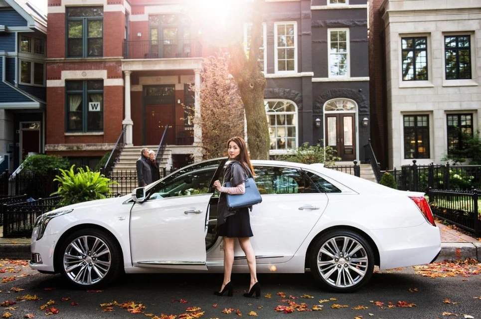 Young woman in black dress exits white Cadillac sedan on city street with brownstone buildings, autumn leaves, and golden sunlight.