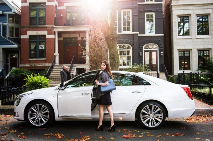 Young woman in black dress exits white Cadillac sedan on city street with brownstone buildings, autumn leaves, and golden sunlight.