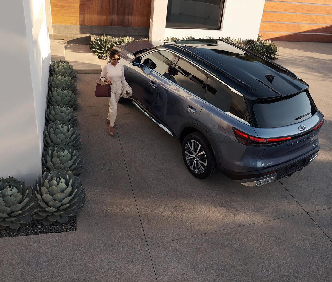 A woman in cream-colored clothing walks next to a blue SUV parked in front of a modern building with desert landscaping.