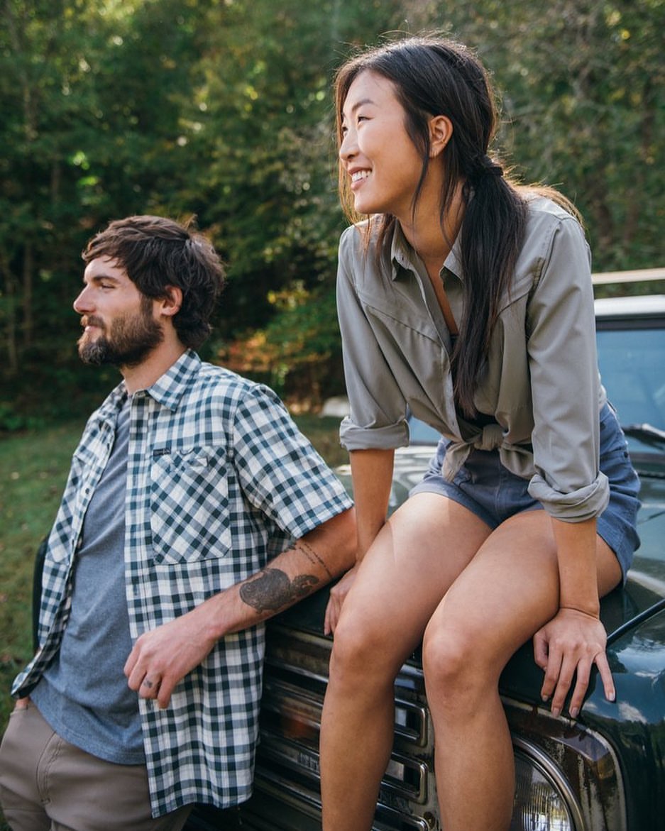 A smiling Asian woman sits on the hood of a dark green SUV next to a bearded man in a plaid shirt in an outdoor wooded setting.