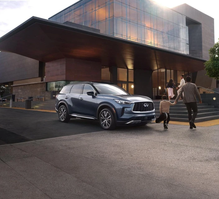 Blue Infiniti QX60 parked near modern building, people walking up stairs. Automotive lifestyle shot with a family. Urban setting, daytime.