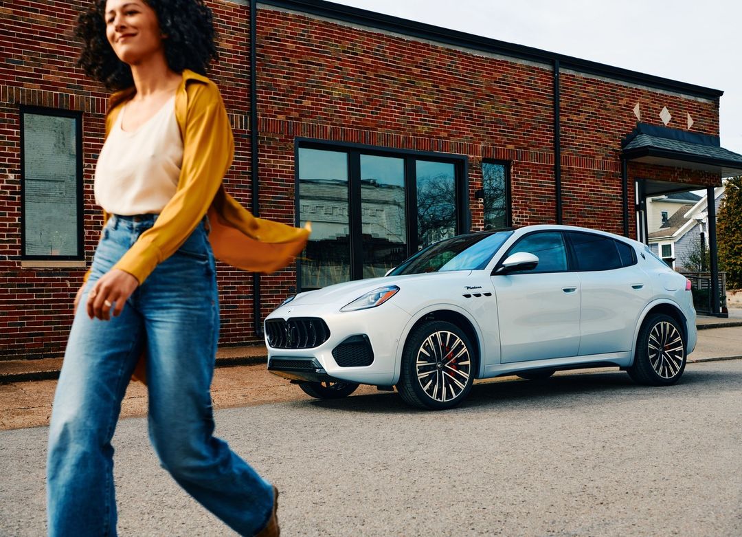 Silver Maserati Grecale SUV parked on a city street with a woman walking by, building in the background, shot in daytime.