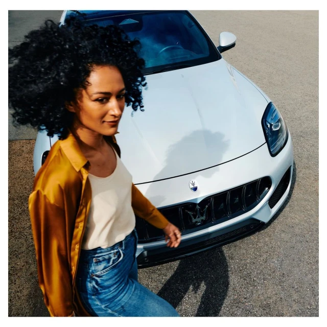 Woman walking near a white Maserati car with black rims on an asphalt surface in daylight, posing with a slight smile.
