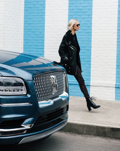 Woman in black jacket and patterned tights leaning on a navy blue Lincoln Navigator parked against a blue and white brick wall.