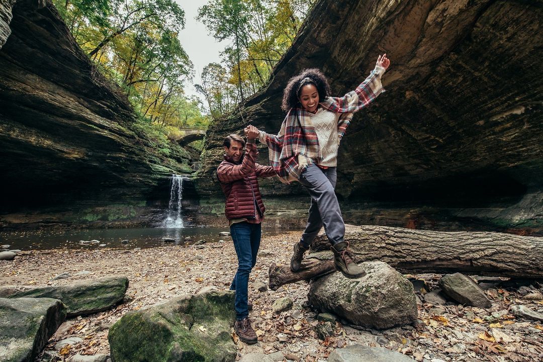 A woman jumps over a log in a riverbed with a man's assistance, surrounded by a rocky canyon, trees, and a waterfall in the background.