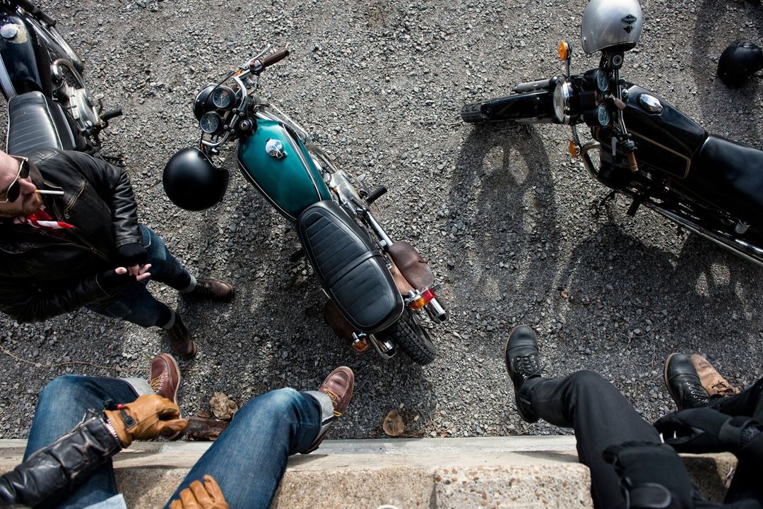 Overhead shot captures three vintage motorcycles parked on gravel, with three people visible from the knees down, showcasing a casual outdoor gathering.