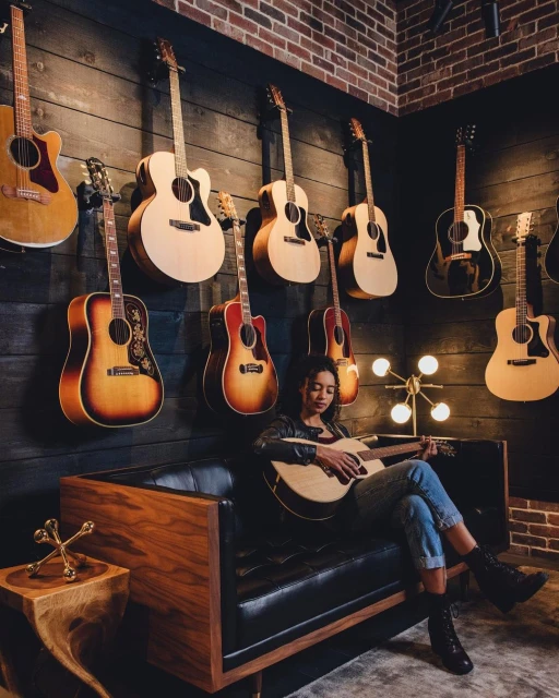 Young woman playing guitar on a black leather couch with acoustic guitars hanging on a wood-paneled wall and brick corner.