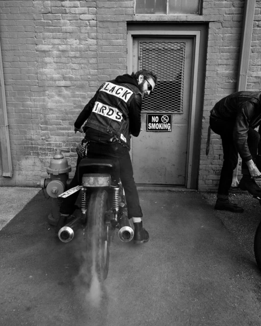Monochrome shot of a motorcycle, with a driver wearing a Black Birds jacket, parked near a brick building and a silver door.