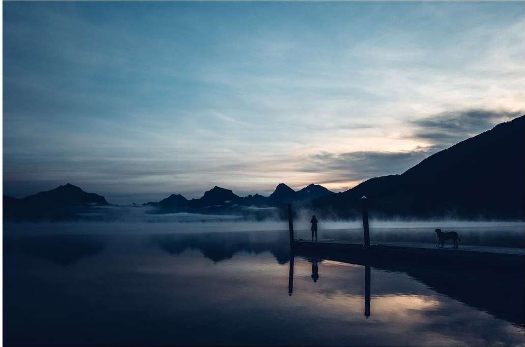 Silhouette of a man and a dog stand on a pier at dusk reflecting on calm water with mountains in the background under a cloudy sky.
