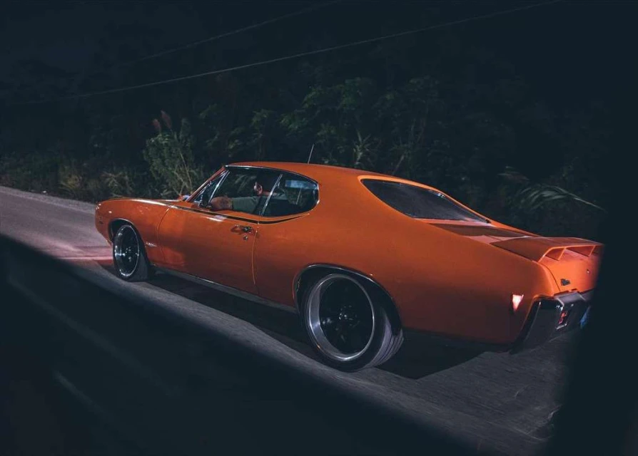 Vintage orange Pontiac GTO driving on a rural road at night. Trees and bushes are visible in the background. 