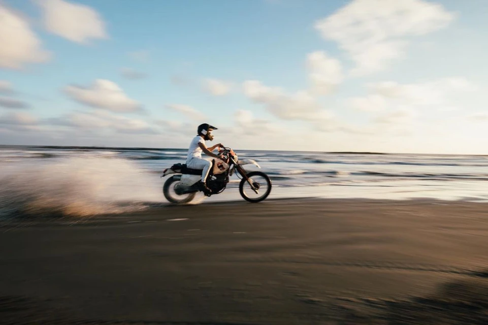 Man riding a dirt bike on a sandy beach with ocean waves, wearing a helmet and casual clothes, at sunset with a cloudy sky.