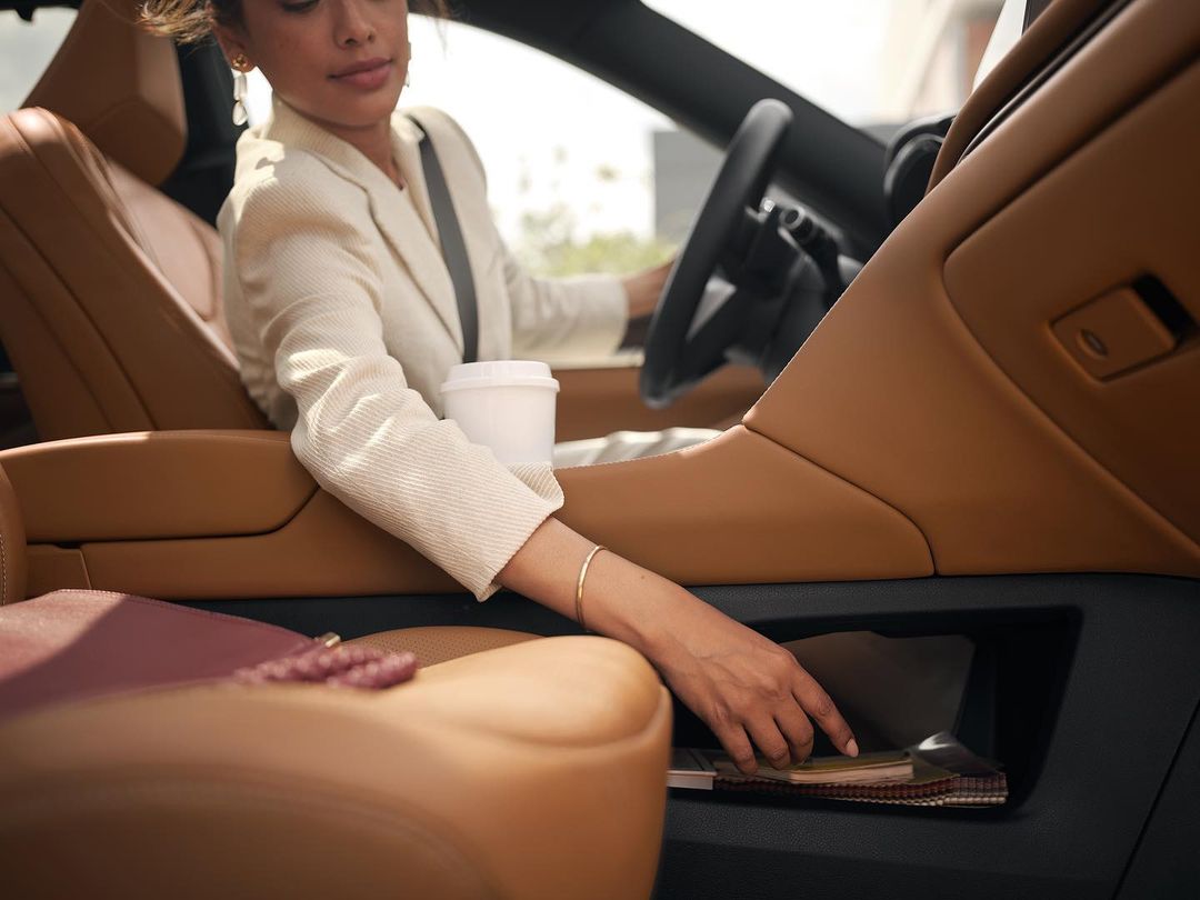 Professional woman driving car in business attire with coffee cup, reaching for items in the side compartment; focus on interior details.